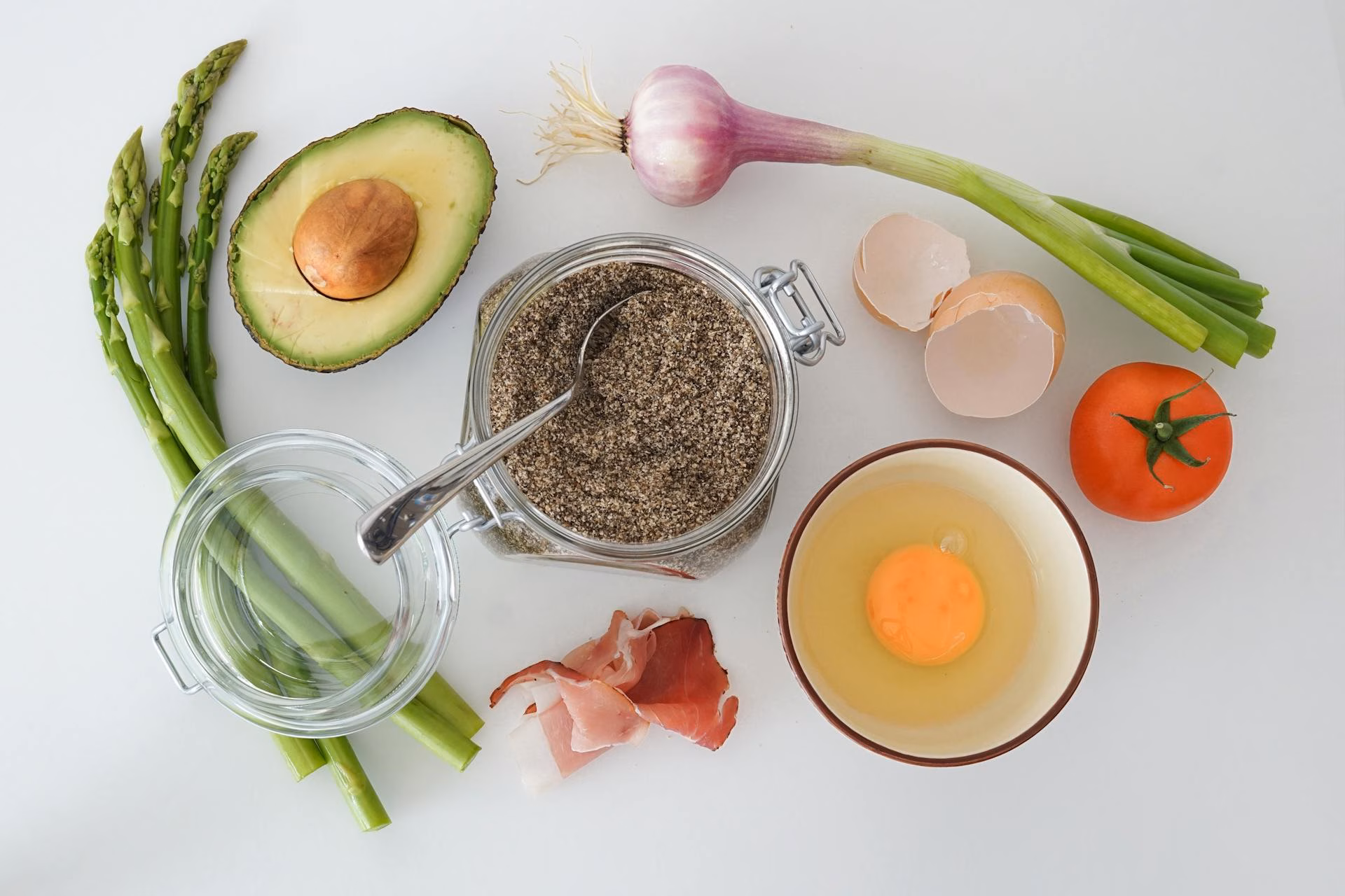 A flat-lay of keto-friendly whole foods including avocado, eggs, tomatoes, herbs and oils